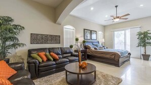 A spacious living area with a black leather sofa, a round coffee table, and a large bed with a headboard. There are two windows with blinds, a ceiling fan, and a large plant in the corner. - Villas House Bayview 3 Bedroom Home by RedAwning (South Padre Island)