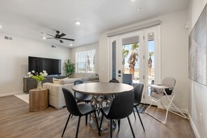 A modern living room with a round wooden table, black chairs, a grey sofa, a flat-screen TV, and a ceiling fan. - Blue Sky Oasis 2 Bedroom Condo (St. George)