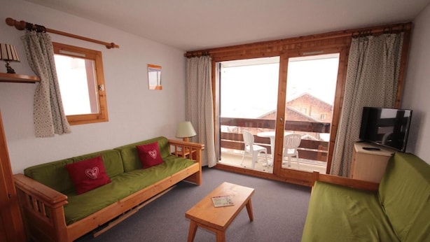 A cozy living room with a green sofa, a wooden coffee table, and a TV on a wooden cabinet. Large windows offer a view of the outside. - 100m from ski slopes, 2nd floor, view ski slopes, balcony, tv, ski locker, 23m², Les Saisies (Les Saisies)