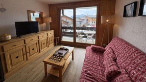 Uma sala de estar aconchegante com um armário de madeira, televisão, sofá vermelho estampado e uma grande janela com vista para edifícios e montanhas.