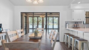 Une cuisine moderne avec des meubles blancs, une table à manger en bois avec des chaises, et une vue sur un lac grâce à de grandes fenêtres.