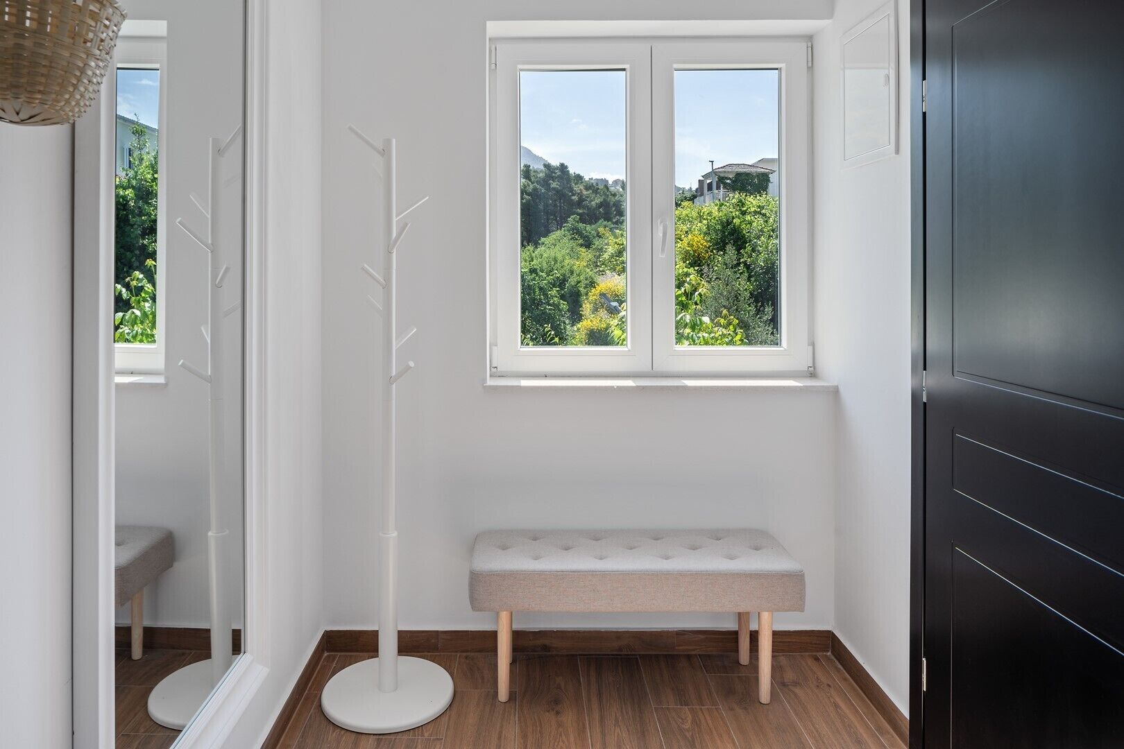 A corner room with a bench, a coat rack, a window with a view of greenery, and a black cabinet.