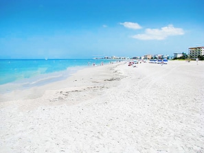 A wide sandy beach with clear blue water, a few people, and buildings in the background.