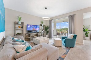 A modern living room with a sofa, ottoman, and a teal armchair. There is a television on a wooden stand, and a large window with a view of the outdoors. - 3171-302 Resort Lake View Disney Universal Orlando (Kissimmee)