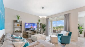A modern living room with a sofa, ottoman, and a teal armchair. There is a television on a wooden stand, and a large window with a view of the outdoors.