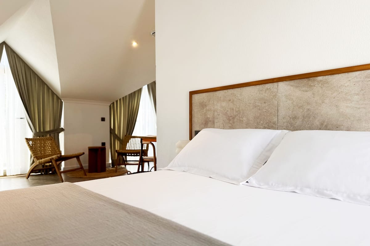 A hotel room with a large bed, a wooden headboard, a small table with chairs, and a view of the outdoors through large windows.