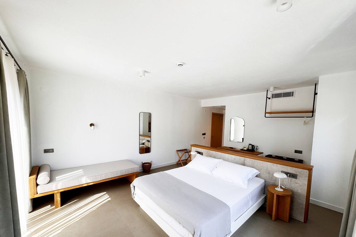 A modern hotel room with a large bed, a small desk, a mirror, and a window with curtains.