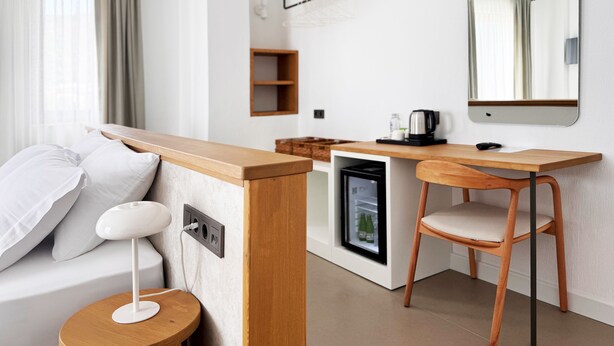 A modern hotel room with a bed, a desk with a chair, a small refrigerator, a mirror, and a window with curtains. - VİLLA KANTARLI (Datça)