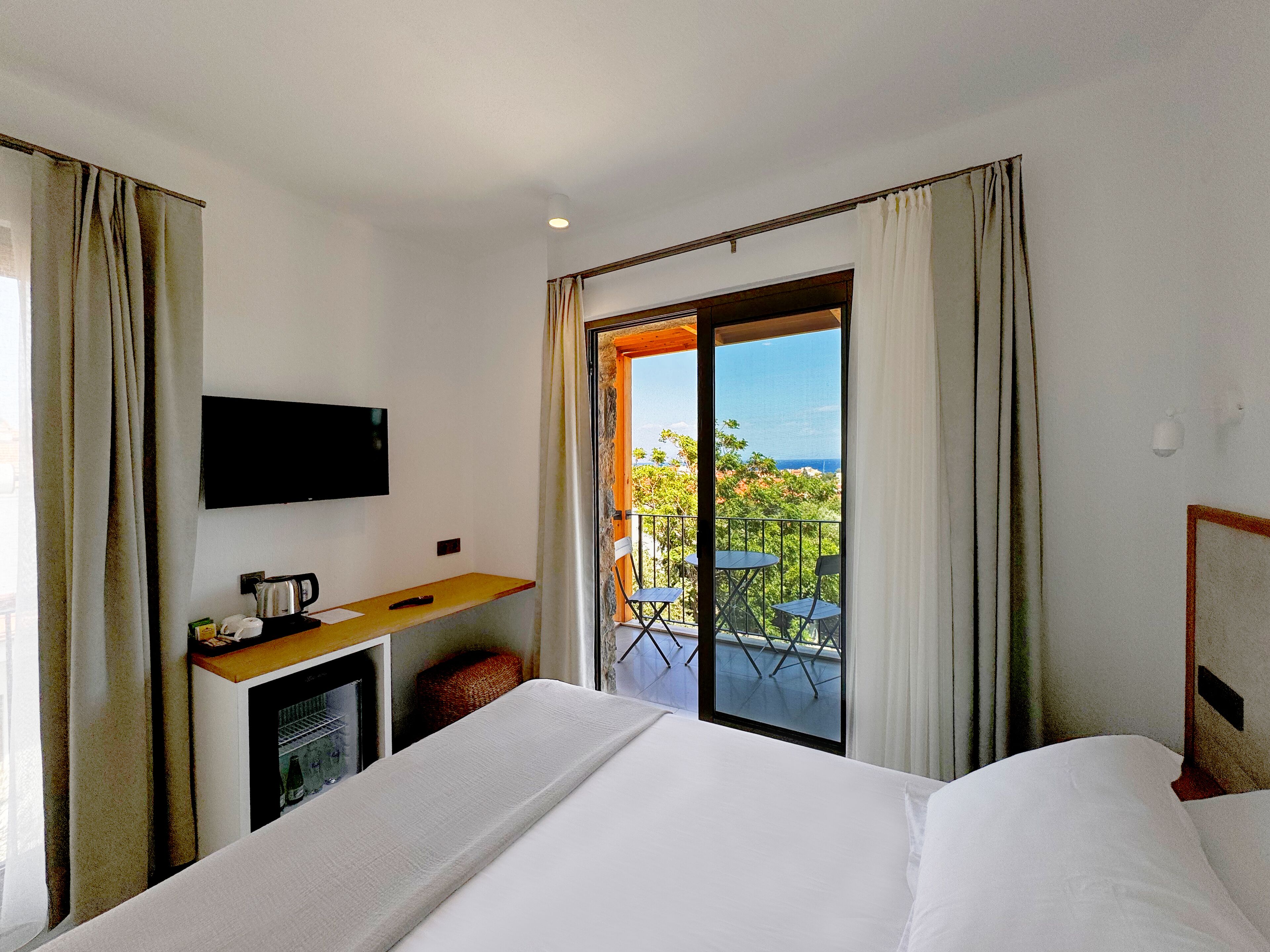A hotel room with a bed, a desk with a TV, a coffee maker, and a view of a balcony with outdoor furniture.