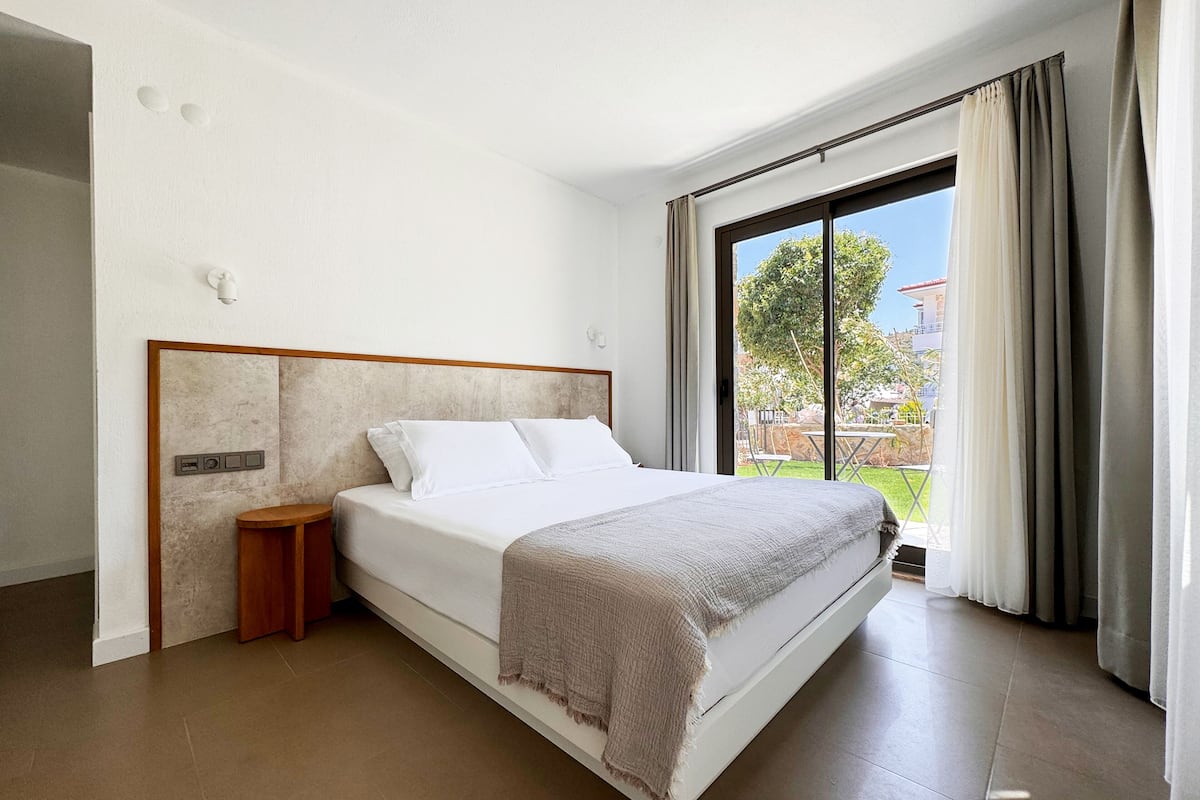 A modern hotel room with a large bed, a wooden nightstand, a wall-mounted control panel, and a large window with curtains.