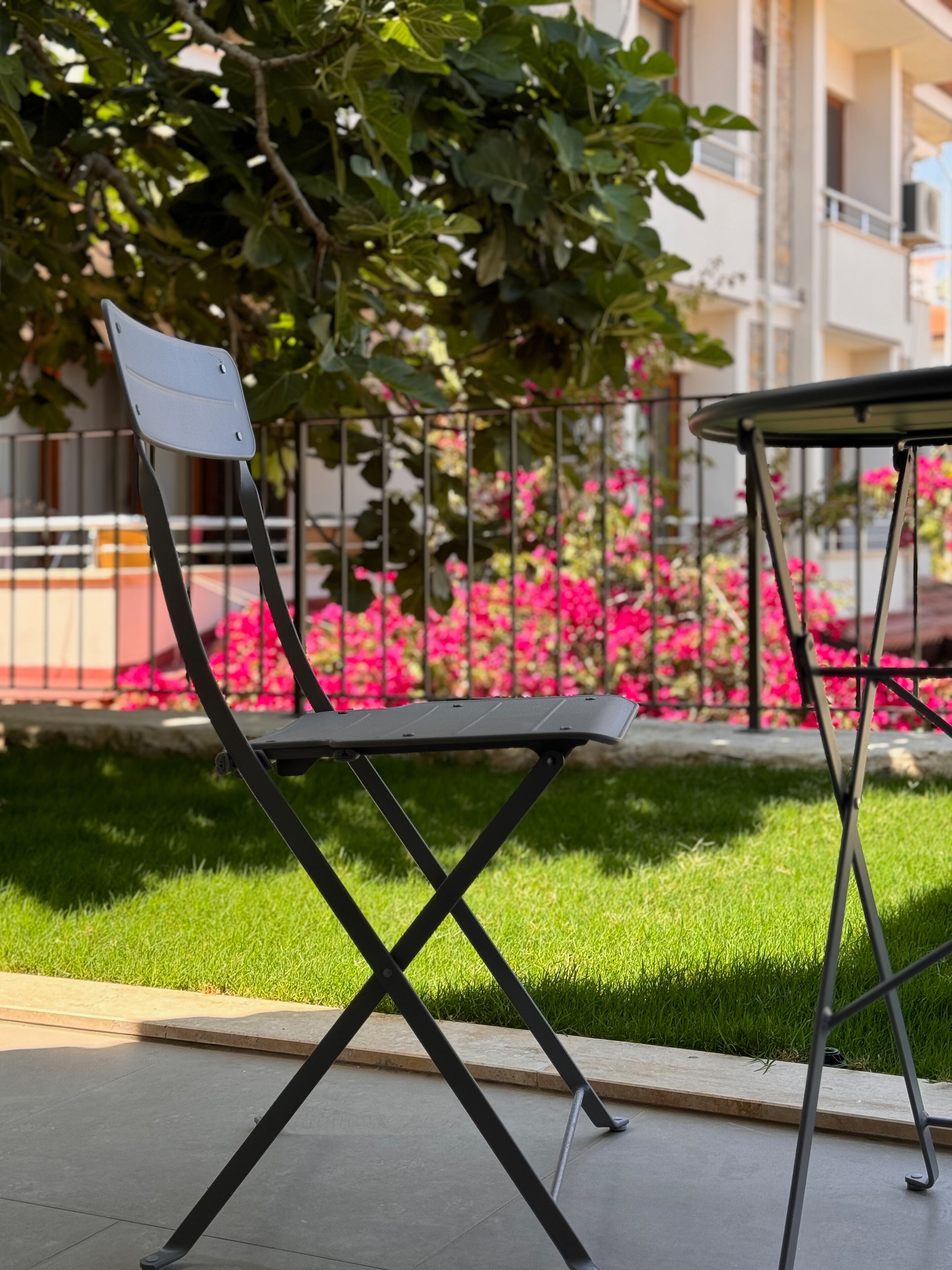 Una silla plegable con estructura metálica y asiento plano, ubicada sobre una superficie pavimentada, con un área de césped y una valla metálica al fondo.