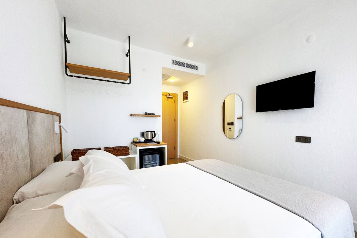 A hotel room with a bed, a TV, a mirror, a shelf with a kettle, and a small table with a lamp.