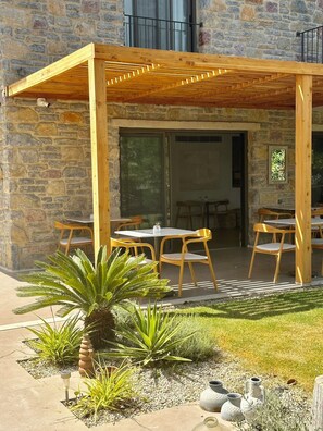 Un área de estar al aire libre techada, con mesas y sillas de madera, un muro de piedra y un pequeño jardín con plantas y macetas decorativas.