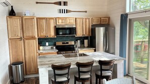A modern kitchen with wooden cabinets, a central island with bar stools, a ceiling fan, and a view of the outdoors through a large window.