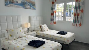 A hotel room with two beds, a nightstand with a lamp, a window with star-patterned curtains, and a beach scene painting on the wall. - Cozy 1 bedroom apartment on a popular complex (Puerto del Carmen)