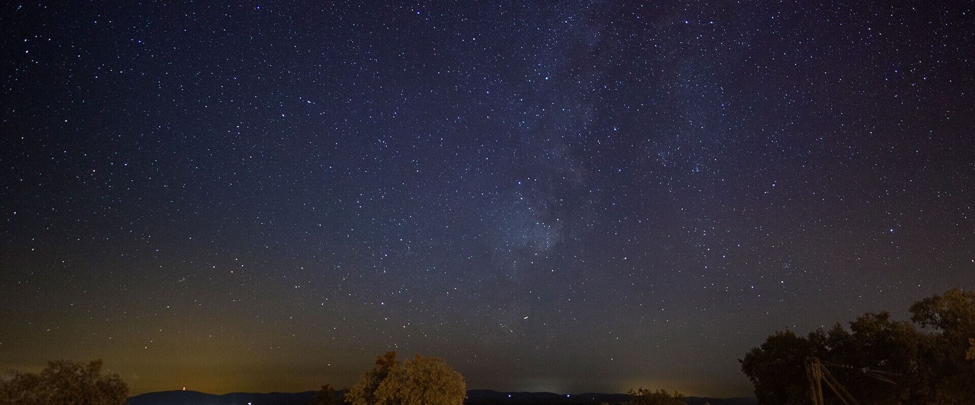 A night sky filled with stars, a tree line, and distant hills.