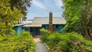 A rustic blue house with a chimney, surrounded by lush greenery and a gravel path leading to the entrance.