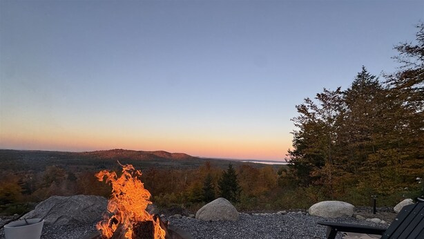 Un foyer avec des bûches, entouré de rochers et agrémenté d’un banc, offrant une vue panoramique sur des collines et un plan d’eau au coucher du soleil.