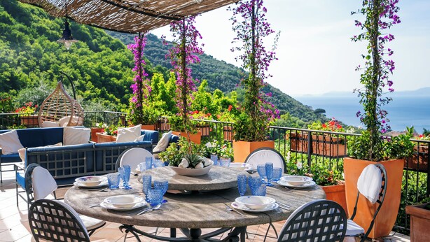 Une table ronde, dressée avec des assiettes, des verres et des couverts, entourée de chaises, sur une terrasse offrant une vue sur un paysage montagneux.