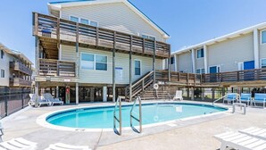 A pool area with a wooden deck, lounge chairs, and a building with multiple levels and balconies.