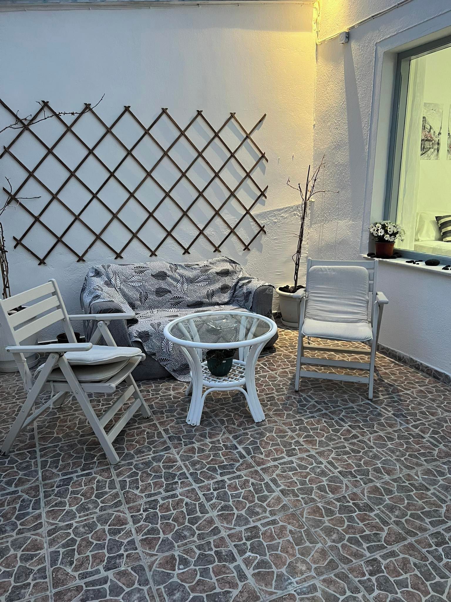 A patio area with a white sofa, a round glass table, and two white chairs. A lattice wall and a window with a view are also visible.