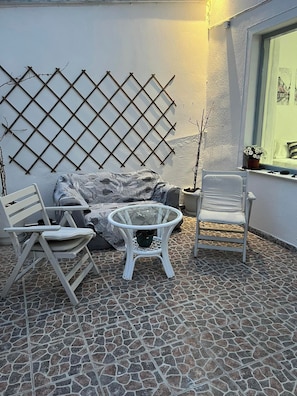 A patio area with a white sofa, a round glass table, and two white chairs. A lattice wall and a window with a view are also visible. - Sagma Beach Apartments next to the beach (Santorini)