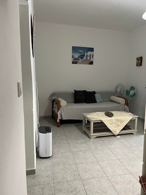 Un salon avec un canapé, une petite table, un ventilateur fixé au mur et une poubelle.