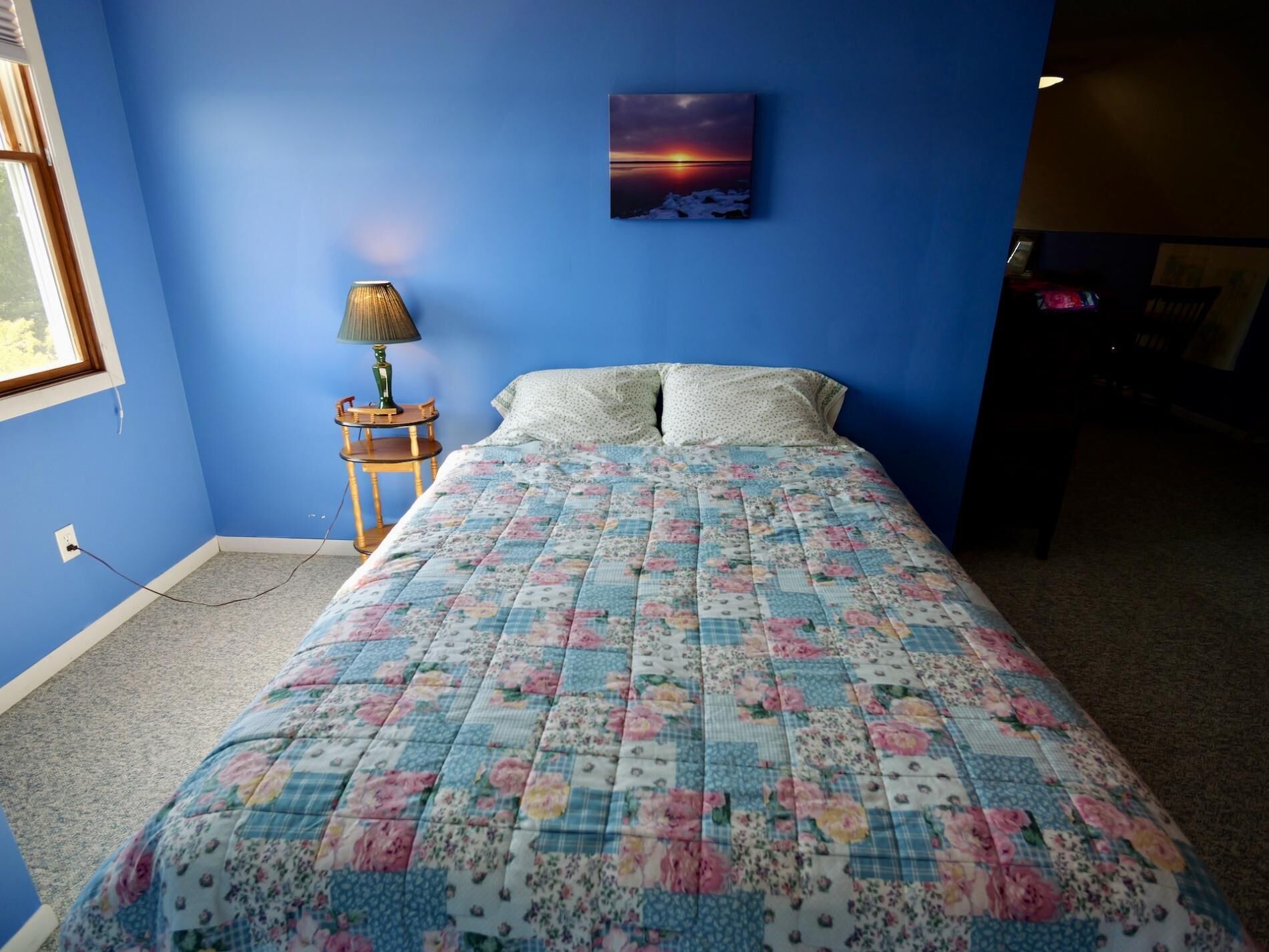 Ein Schlafzimmer mit einem floralen gesteppten Bettbezug, einem kleinen Nachttisch mit Lampe und einem Sonnenuntergangsbild an der Wand.