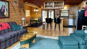A cozy cabin interior with a wooden dining area, a kitchen with a black refrigerator, and a living room with a plaid sofa and a wooden coffee table.