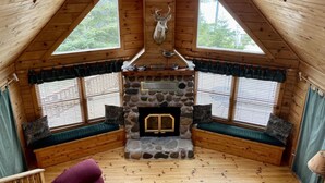 A cozy cabin interior with a stone fireplace, wooden beams, and a large window with a view of trees.