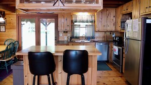A wooden cabin kitchen with a central island, two black bar stools, a stainless steel refrigerator, and a sink.