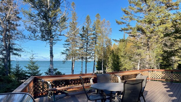 A deck with a table and chairs, overlooking a body of water and surrounded by trees.