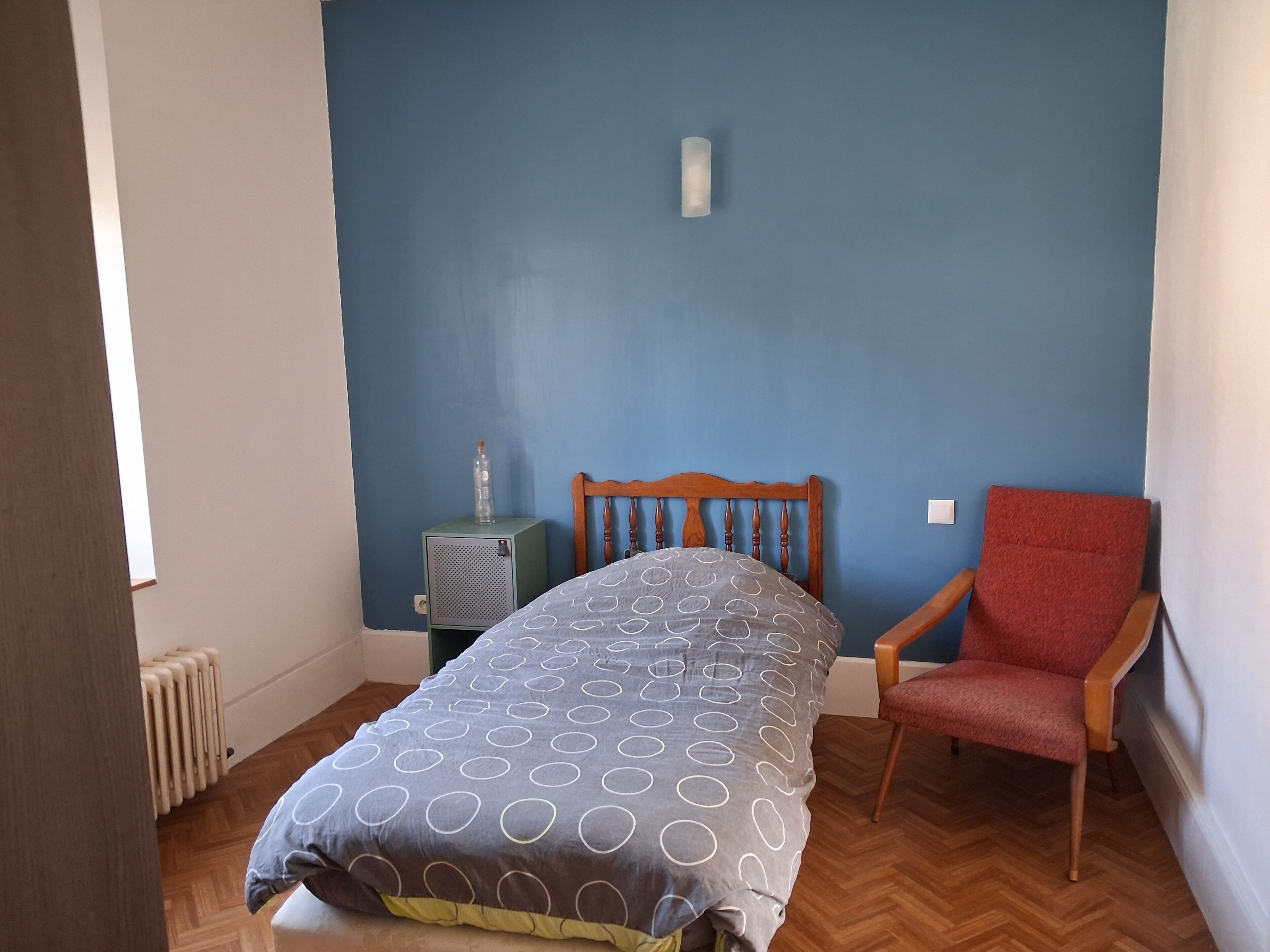 A bedroom with a bed, a chair, a radiator, and a wall with a light switch.