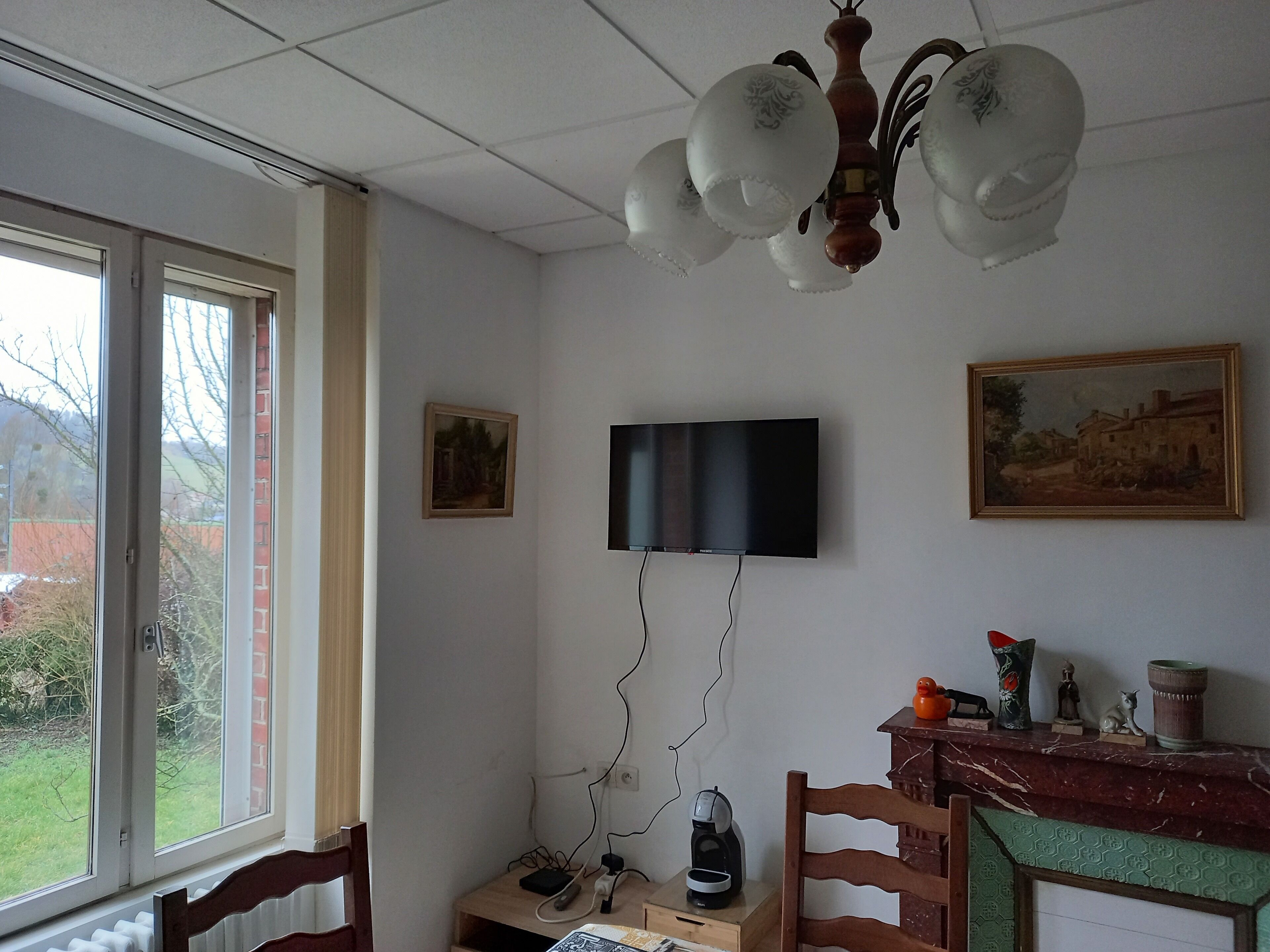 Una habitación con una televisión montada en la pared, una silla de madera, una mesita con un hervidor y una ventana que da a vistas de vegetación.