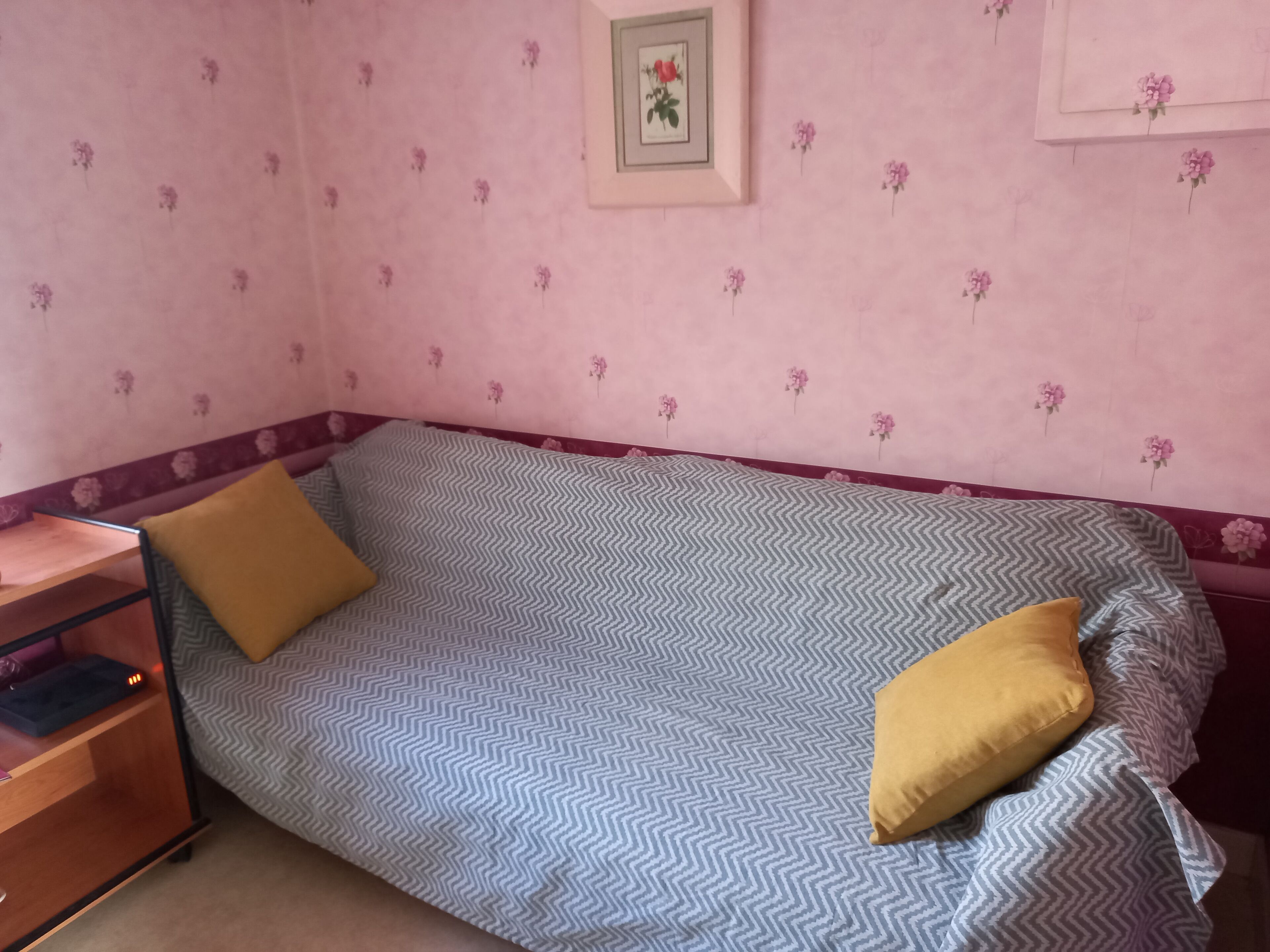 A small, single-bed room with a chevron-patterned bedspread, a wooden nightstand, and a framed picture on the wall.