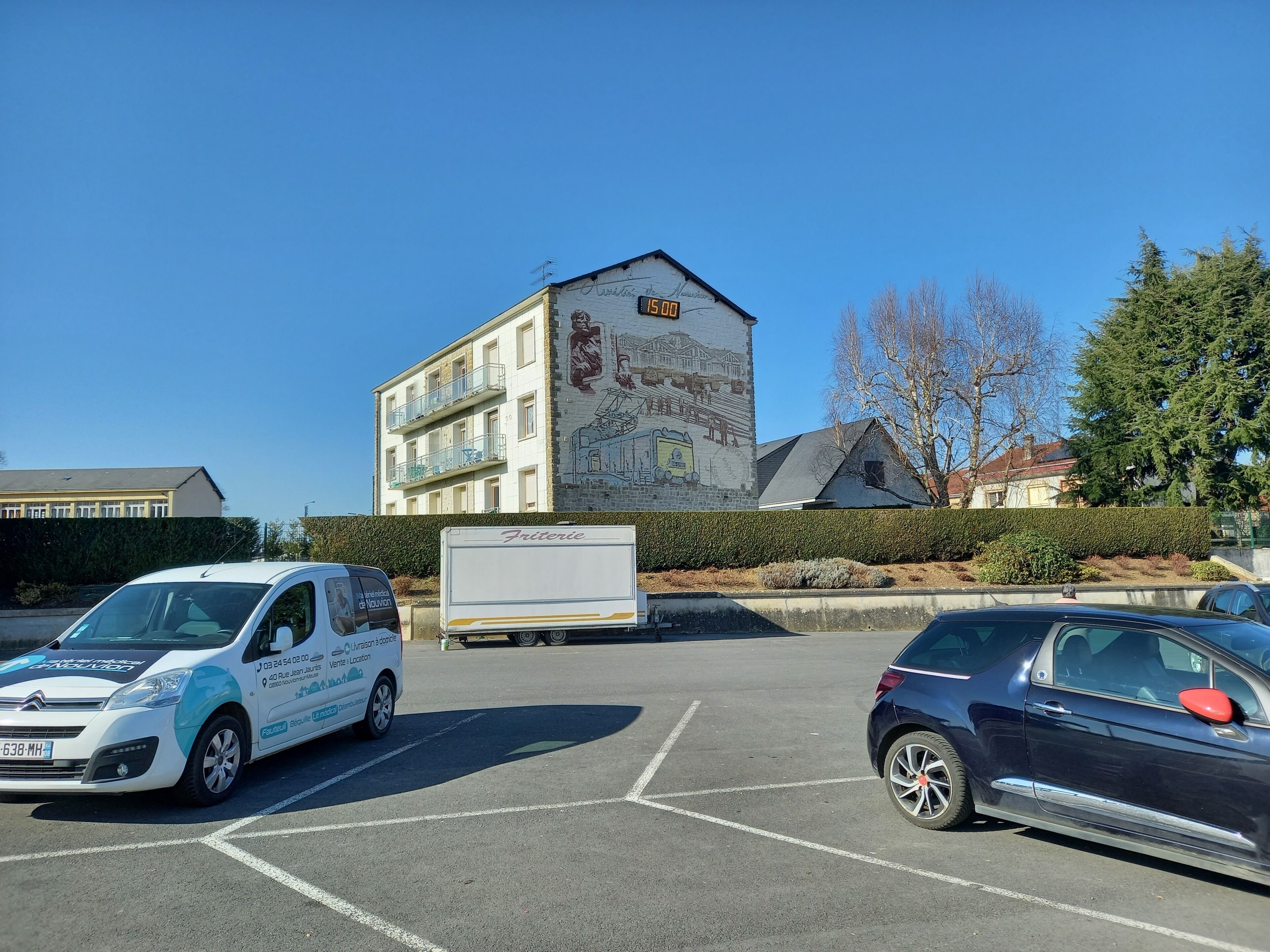 Een parkeerplaats met een witte bestelwagen en een blauwe auto. Op de achtergrond staat een gebouw met een muurschildering en een klok waarop 15:00 uur staat.
