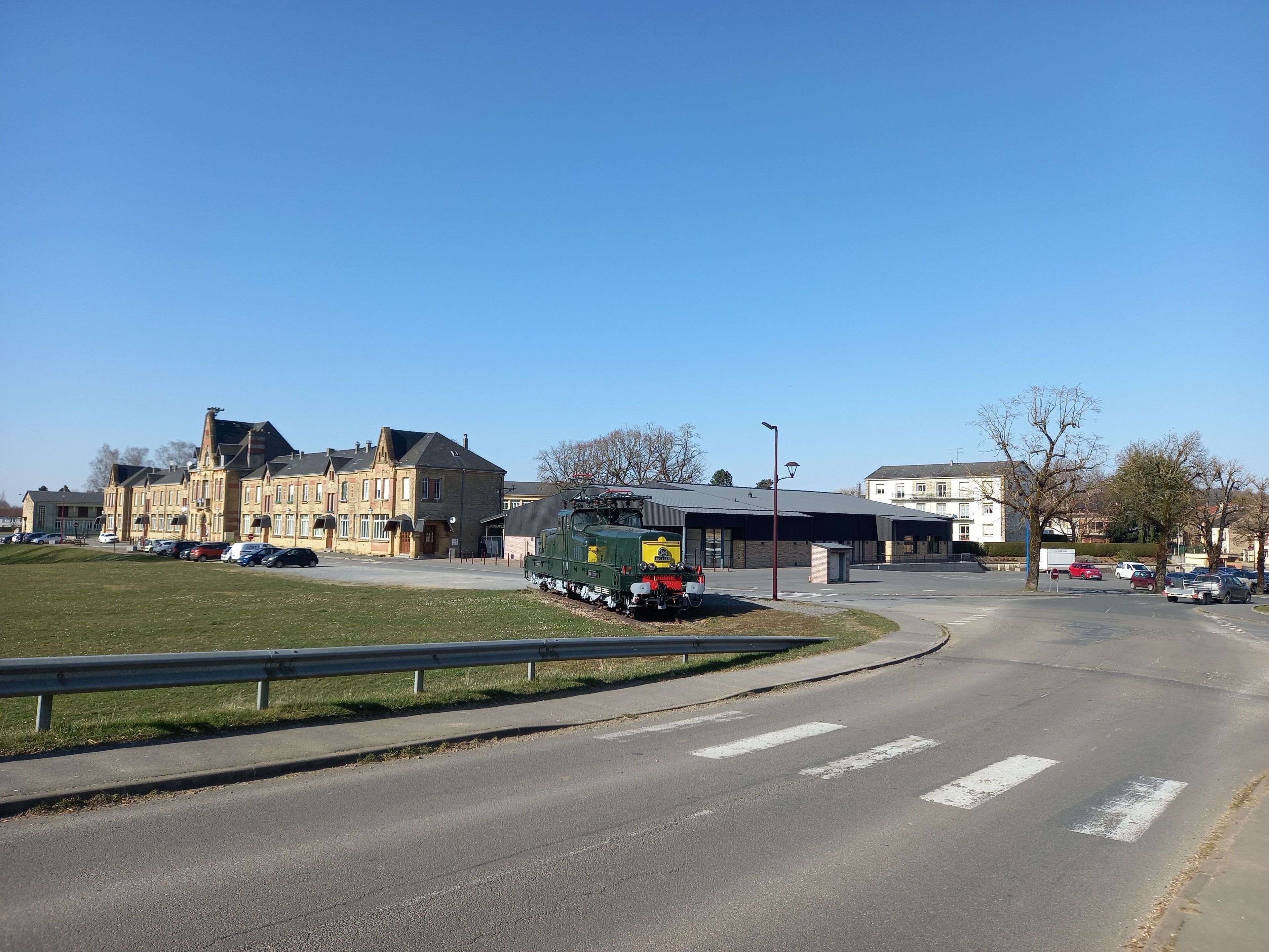 A train station with a green locomotive, a parking lot with cars, and a building with a large entrance.