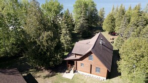 A cabin with a sloped roof, surrounded by dense forest and a lake.