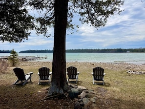 Paisagem à beira de um lago, com quatro cadeiras Adirondack sob uma grande árvore, margem rochosa e um píer que se estende sobre a água.