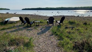 Ein Kiesstrand mit vier Adirondack-Stühlen und einem kleinen Tisch mit Blick auf einen ruhigen See und eine bewaldete Uferlinie.