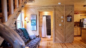 Ein gemütliches Blockhaus-Interieur mit Holzboden, einer Couch mit gemusterten Kissen, einer Lampe, einem Willkommensschild und einer Küchenzeile.