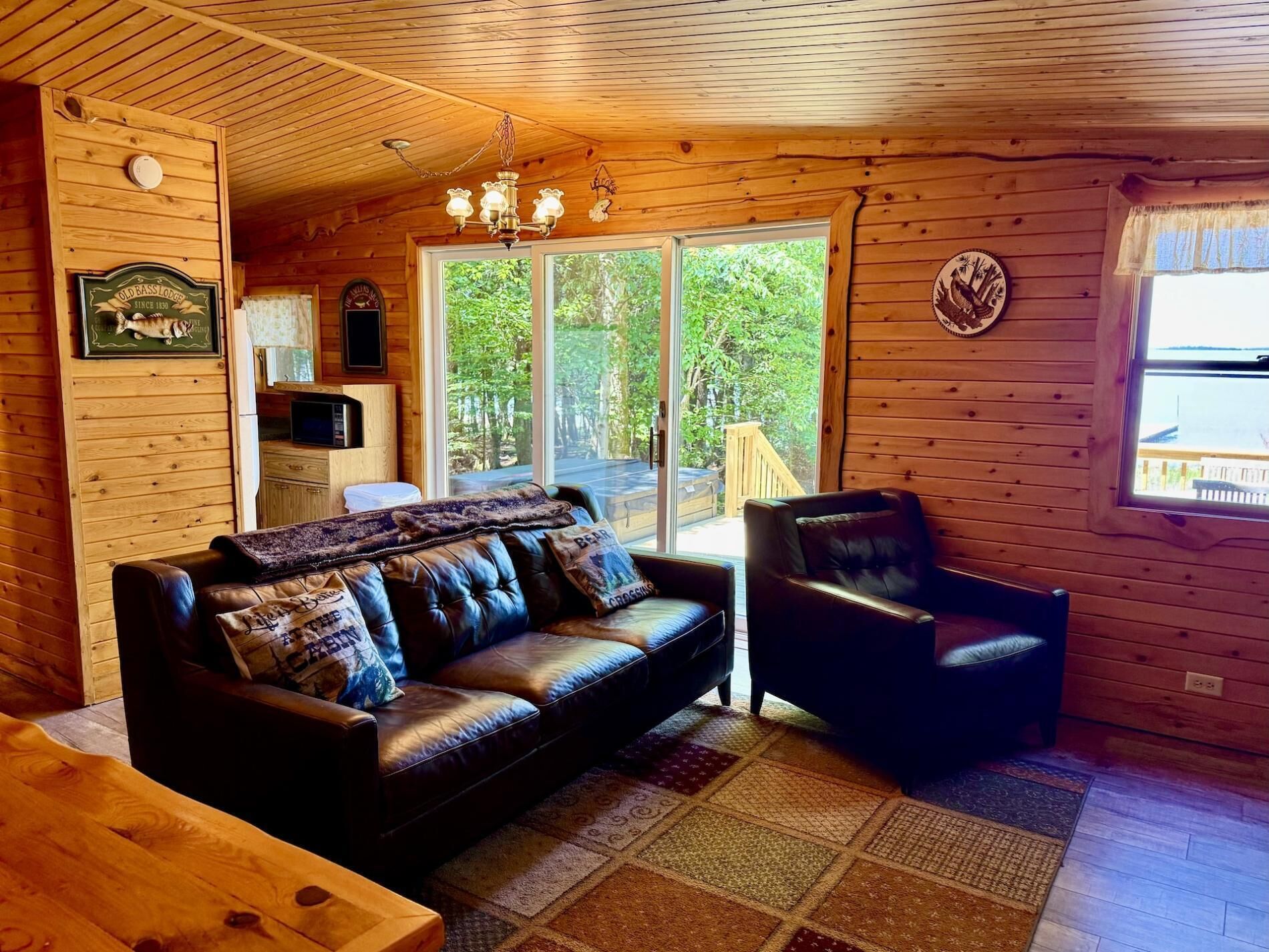 A wooden cabin interior with a leather sofa, a chair, a wooden table, and a large window offering a view of the outdoors.