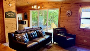 Interior de una cabaña de madera con un sofá de cuero, una silla, una mesa de madera y un ventanal con vistas al exterior.