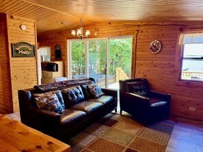 Ein Holzblockbau-Innenraum mit Ledersofa, Stuhl, Holztisch und einem großen Fenster mit Blick ins Freie.
