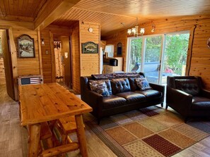 Ein Holzblockhaus im Inneren mit einem Holztisch, einem Ledersofa und einem großen Fenster.