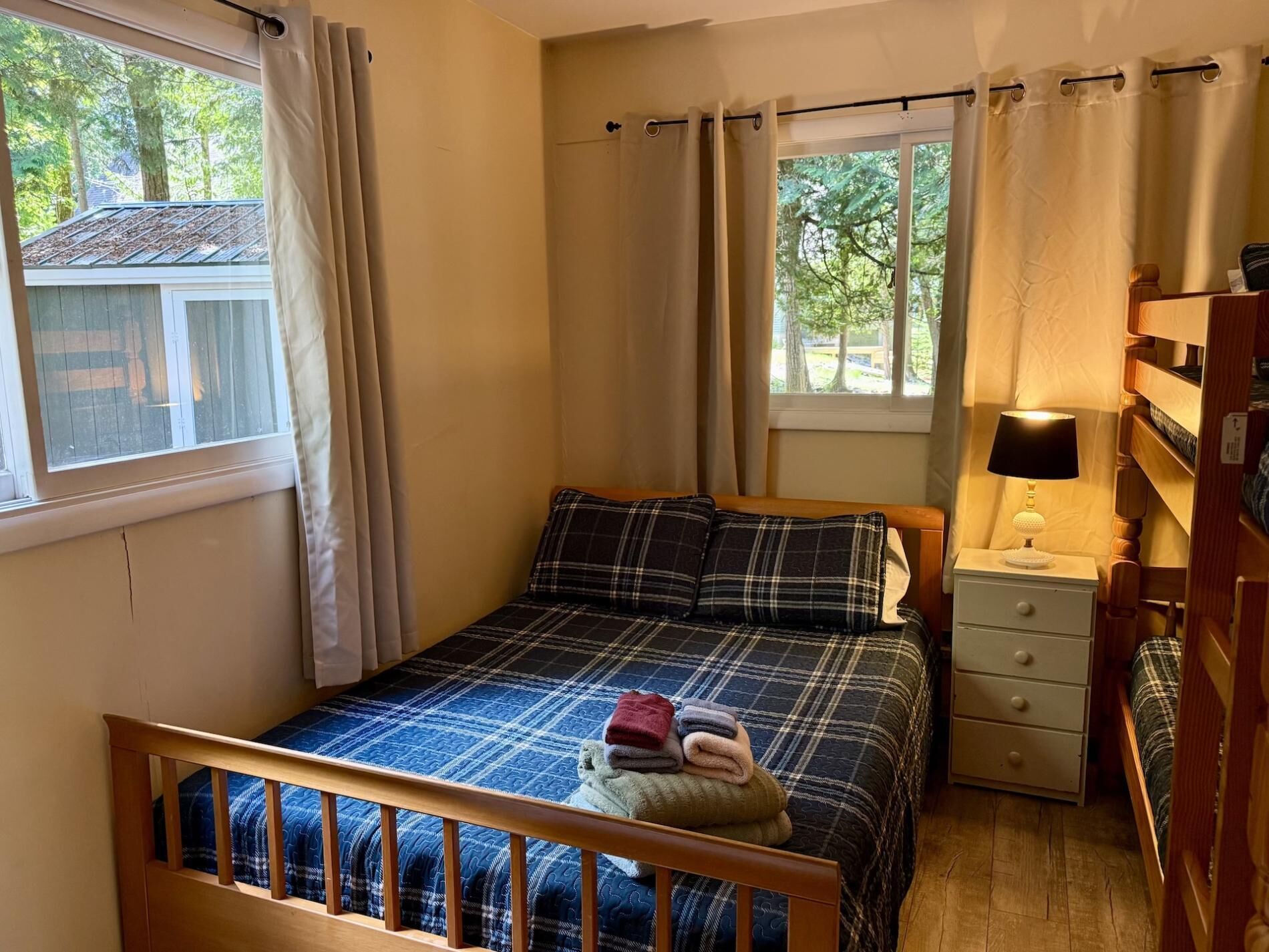Un dormitorio con una cama, una mesita de noche, una lámpara, un ventanal con cortinas y vistas a árboles.