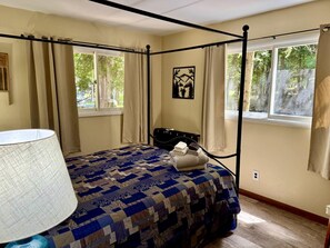 Una cama con dosel, un edredón estampado, una lámpara de noche, una mesita de noche con lámpara y una ventana con cortinas.