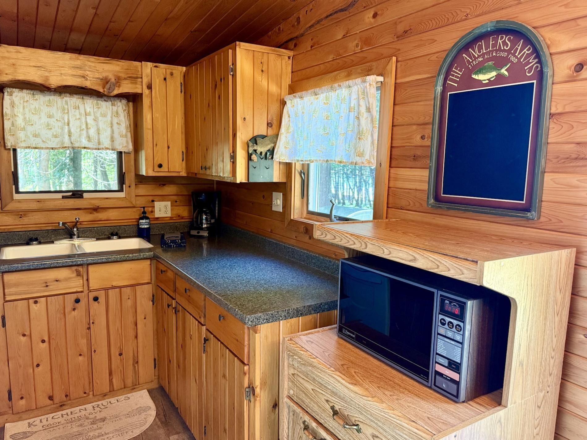 A wooden cabin kitchen with a sink, cabinets, a microwave, and a window with a sign reading 'The Anglers Arms'.