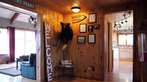 Un interior de madera con un letrero que dice 'WELCOME', una cabeza de oso montada en la pared, documentos enmarcados y una cocina visible a través de una puerta abierta.