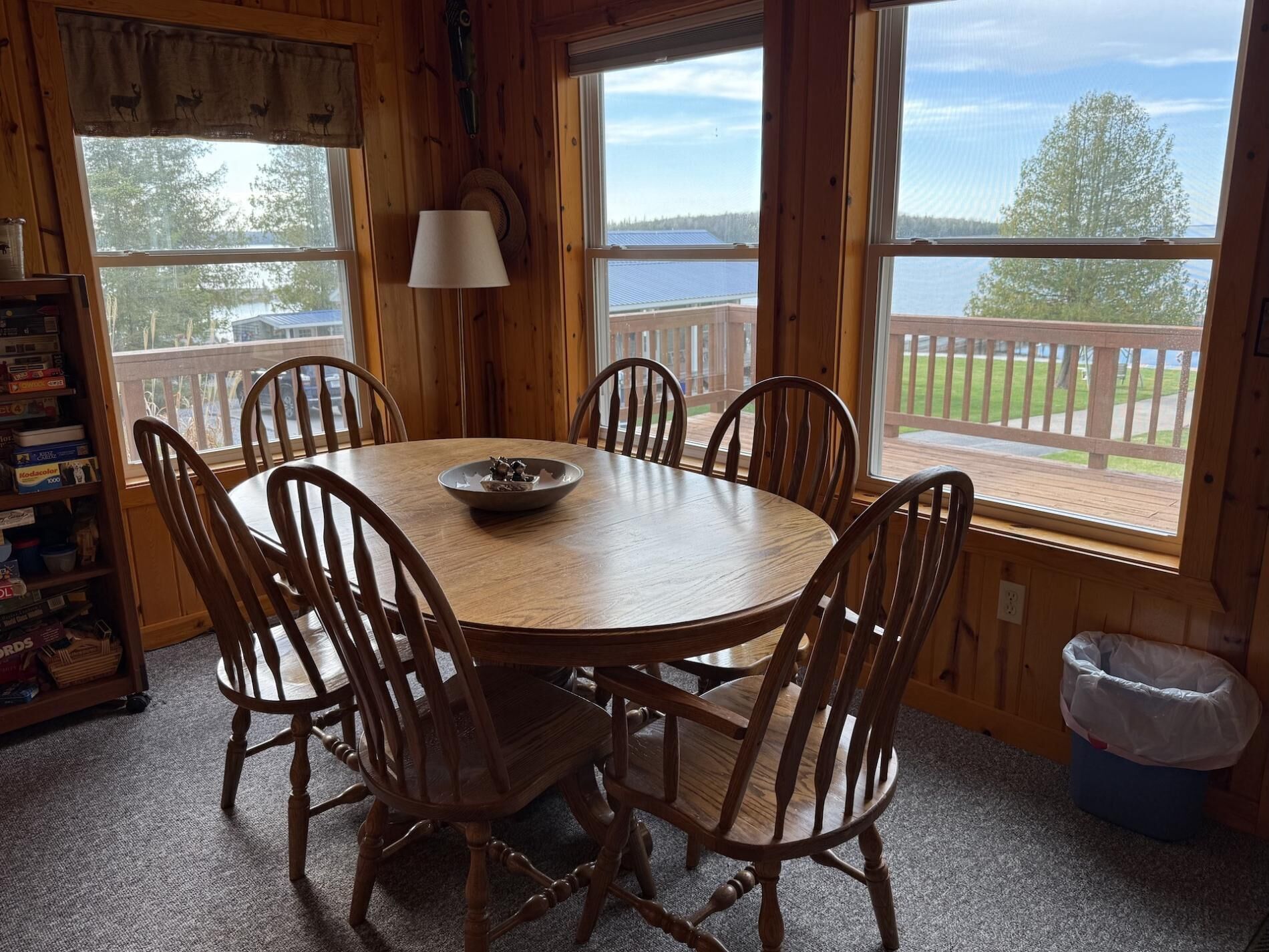 A wooden dining room with a round table and six chairs, large windows with a view of a deck and trees, and a lamp on the wall.